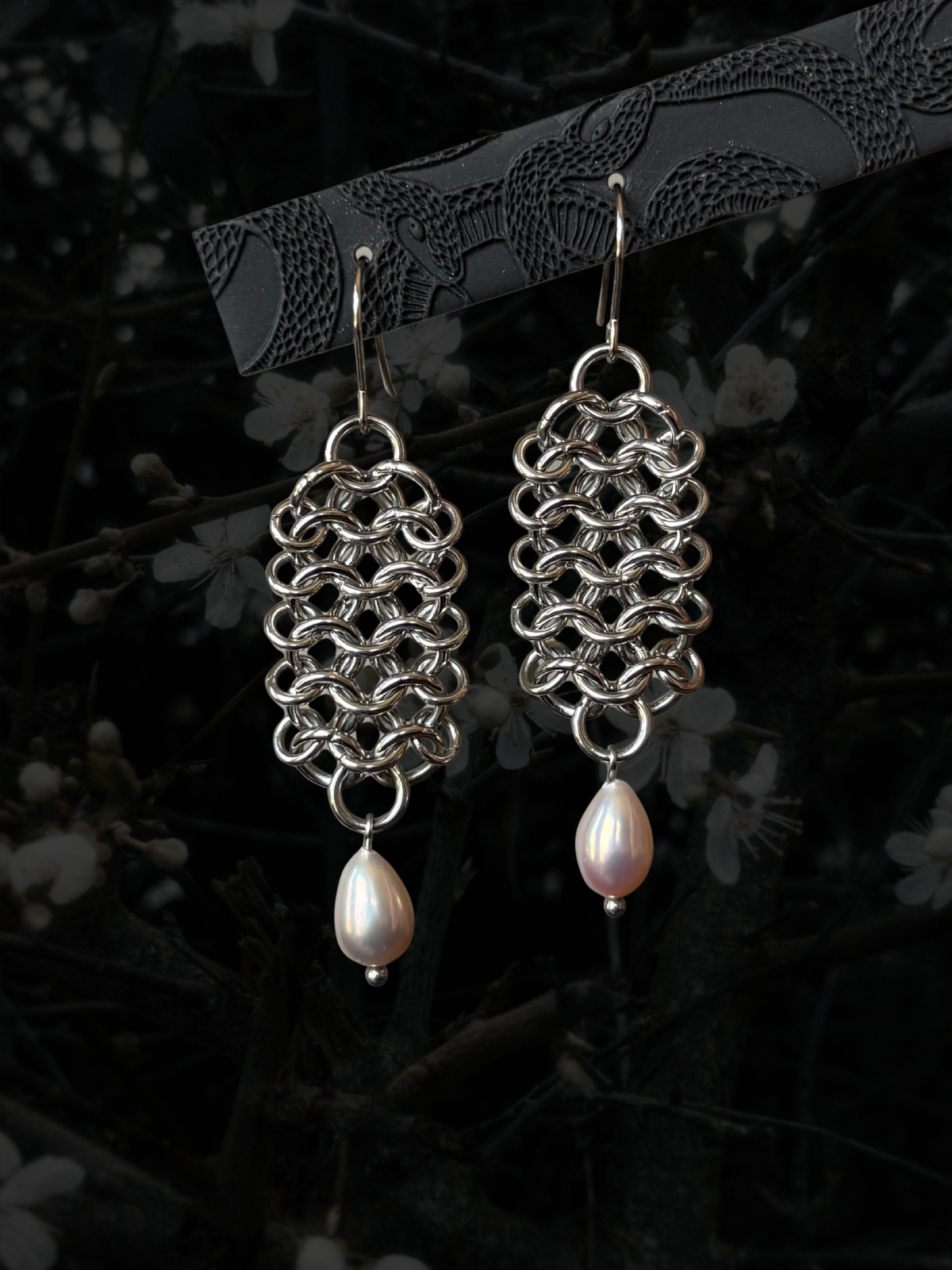 A pair of handmade silver-tone chainmail earrings with teardrop-shaped white freshwater pearls hanging from silver fishhook ear wires, displayed against a dark, blurred background of branches and white blossoms.