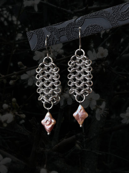 A pair of handmade dangle earrings with silver-toned chainmail loops and irregular pink baroque pearl drops, hanging from a black branch against a dark, blurred background of white blossoms.