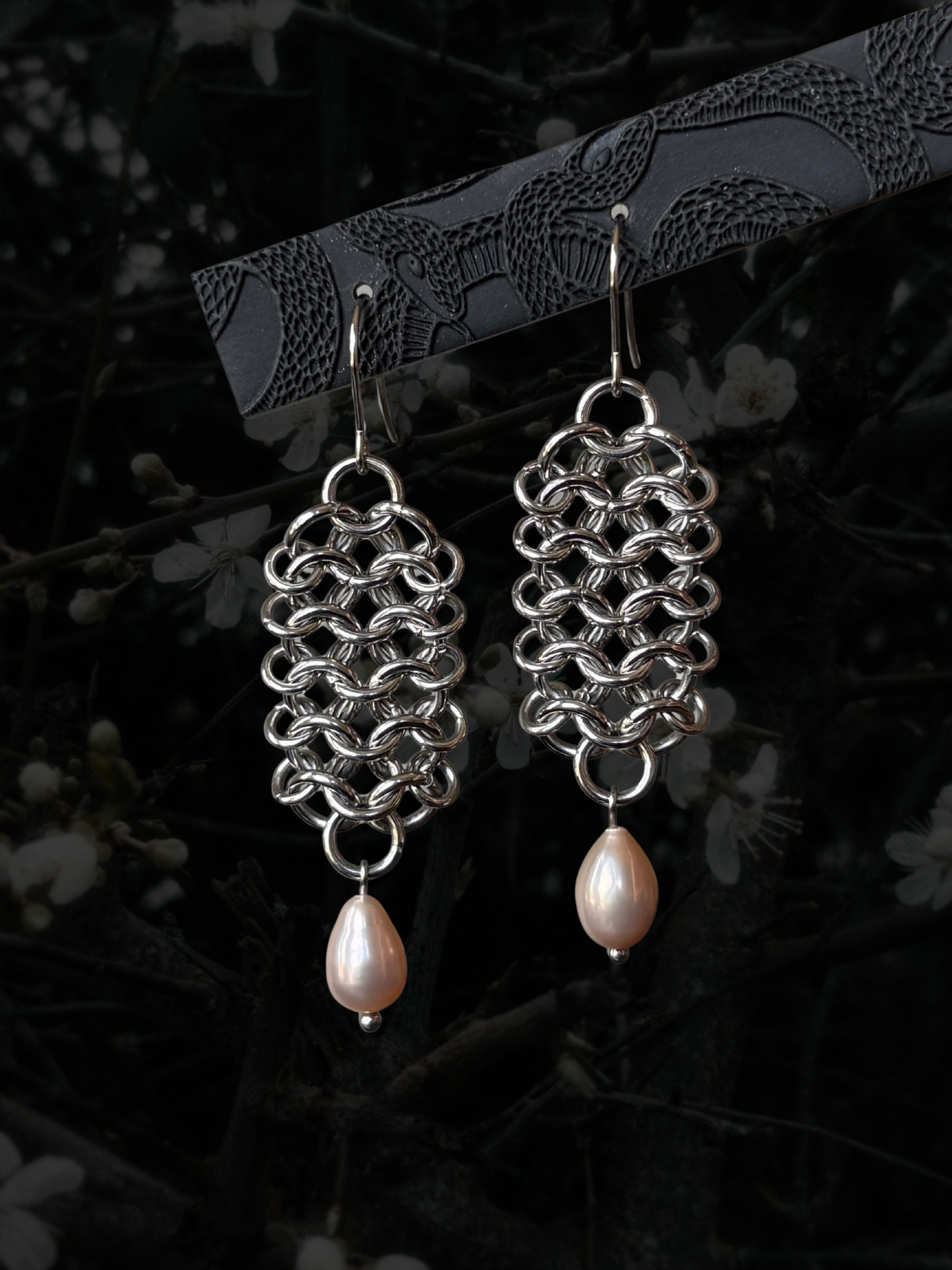A pair of silver chainmail-style dangle earrings with teardrop-shaped pale pink pearl drops, hanging from a dark branch against a dark, blurred background with faint white blossoms.