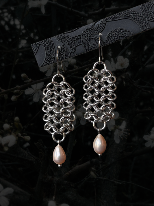 A pair of silver chainmail-style dangle earrings with teardrop-shaped pale pink pearl drops, hanging from a dark branch against a dark, blurred background with faint white blossoms.