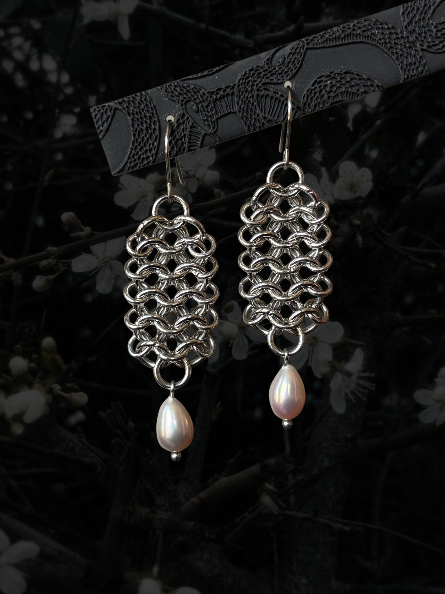 A pair of handmade silver-tone chainmail earrings with teardrop-shaped white freshwater pearls hanging from silver fishhook ear wires, displayed against a dark, blurred background of branches and white blossoms.