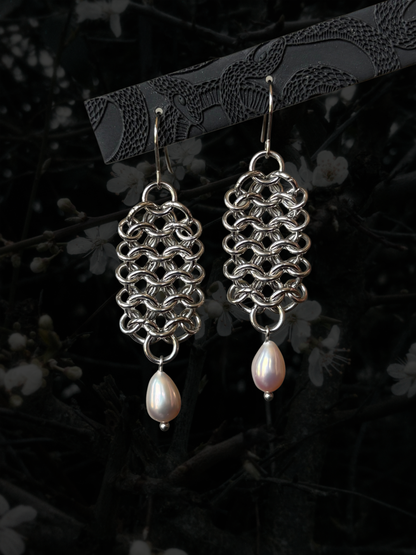 A pair of handmade silver-tone chainmail earrings with teardrop-shaped white freshwater pearls hanging from silver fishhook ear wires, displayed against a dark, blurred background of branches and white blossoms.