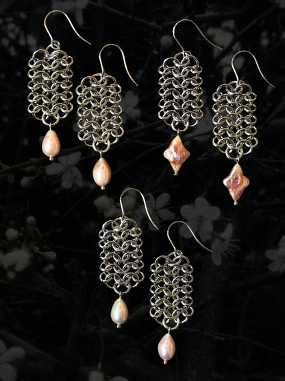 Three pairs of handmade silver-tone chainmail earrings with dangling pink freshwater pearl beads, displayed against a dark, blurred background of white blossoms.