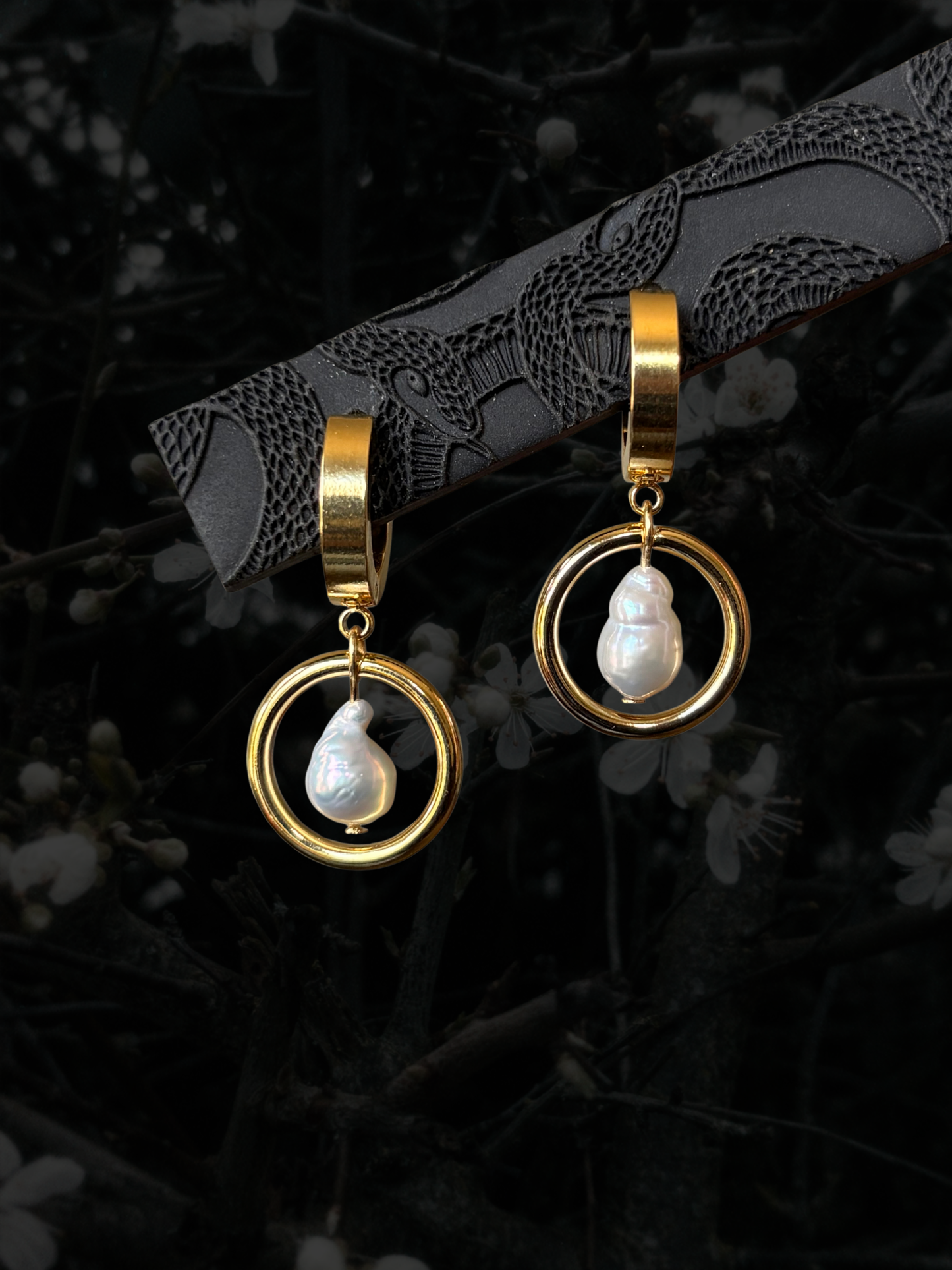 A pair of gold hoop dangle earrings with white teardrop-shaped freshwater pearls, displayed on a dark textured branch against a dark, blurred background of branches and small white flowers.