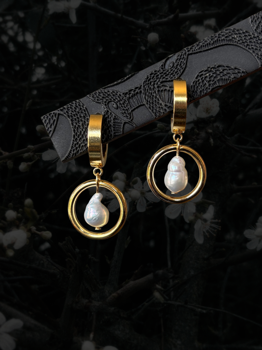 A pair of gold hoop dangle earrings with white teardrop-shaped freshwater pearls, displayed on a dark textured branch against a dark, blurred background of branches and small white flowers.