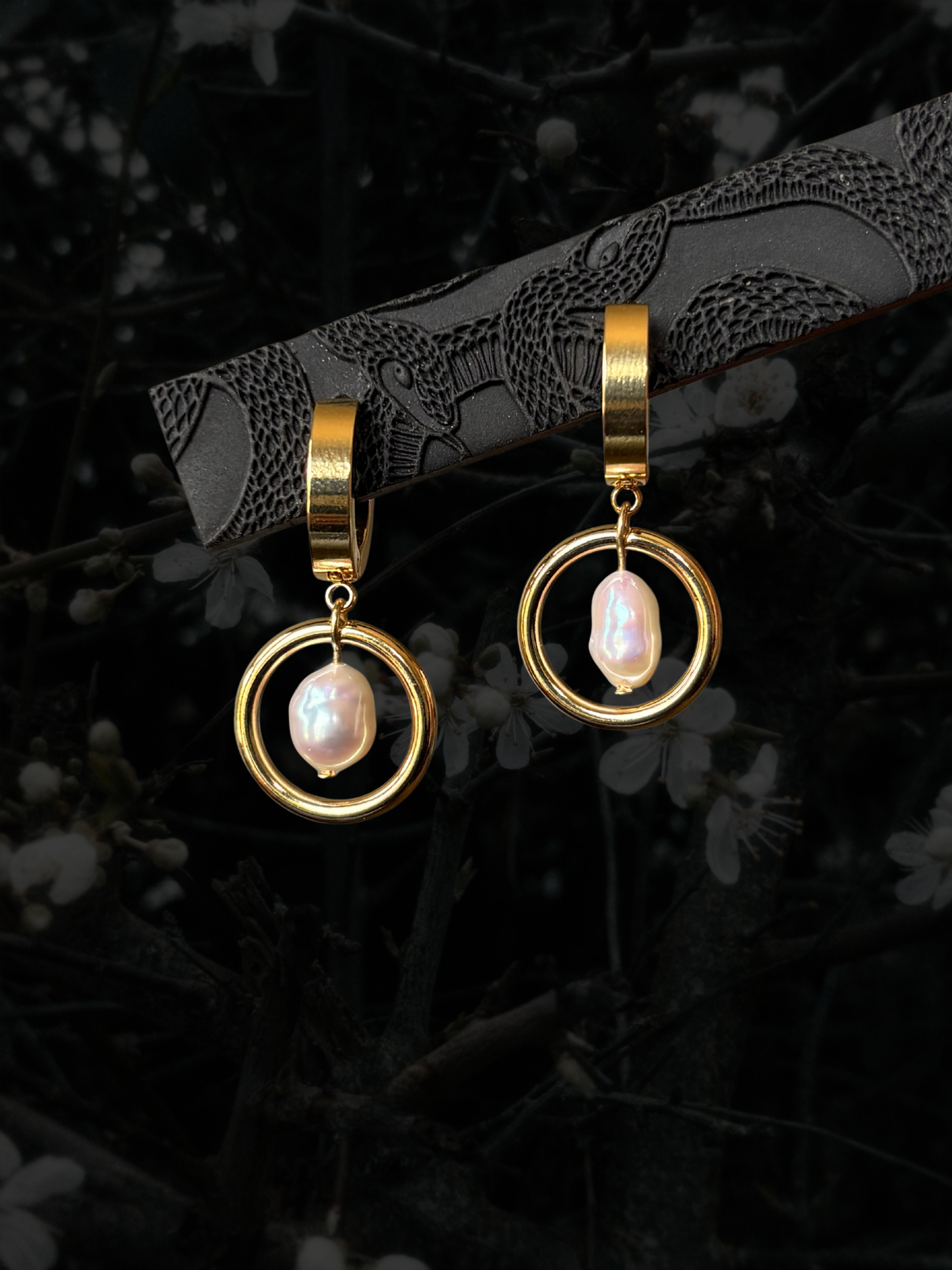 A pair of gold hoop dangle earrings with irregular white baroque pearls, displayed on a dark branch with a black background of blurred flowers.