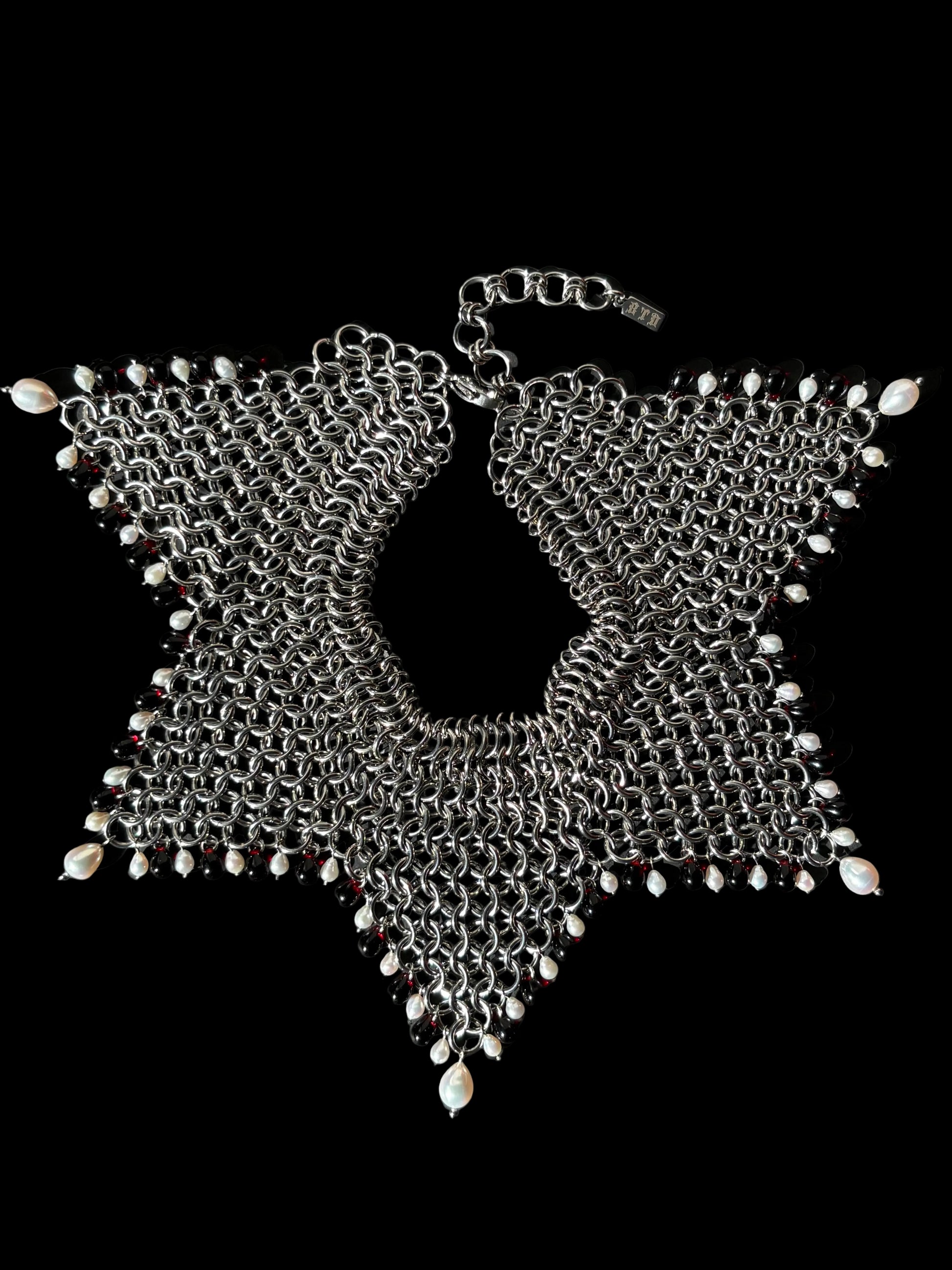A stainless steel chainmail collar necklace with red glass beads and white teardrop pearls, laid flat on a black background.
