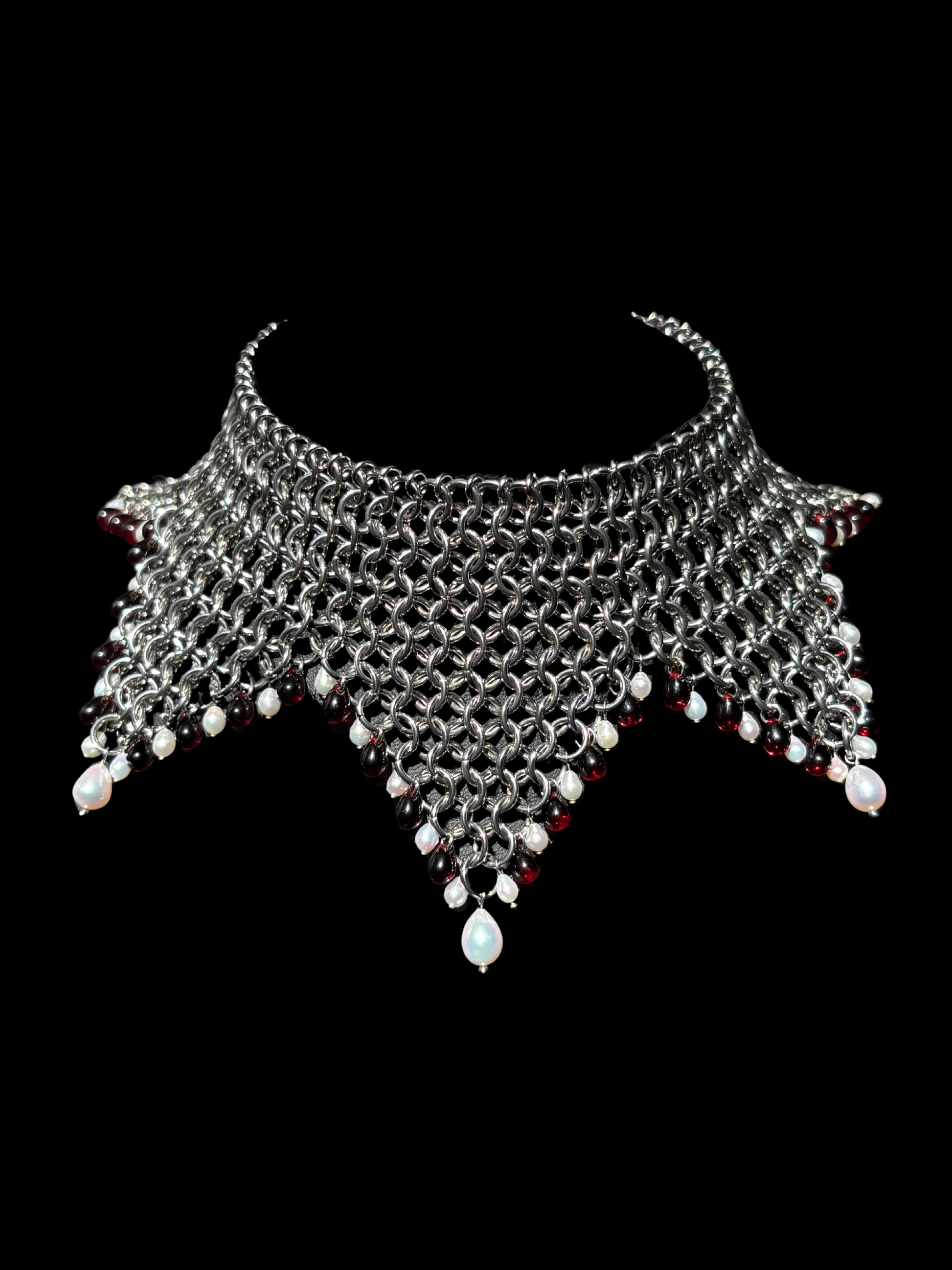 A stainless steel chainmail jester collar necklace with dangling white pearls and red glass beads, displayed against a solid black background.