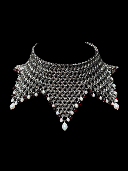 A stainless steel chainmail jester collar necklace with dangling white pearls and red glass beads, displayed against a solid black background.